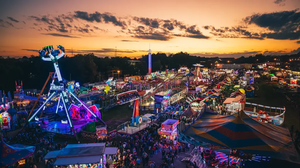 Vista aérea al atardecer de una feria con atracciones iluminadas y ambiente festivo.