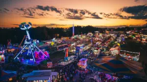 Vista aérea al atardecer de una feria con atracciones iluminadas y ambiente festivo.