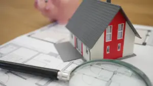 Maqueta de casa junto a una lupa sobre planos arquitectónicos y una hucha.