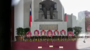 Una estatua conmemorativa rodeada de ofrendas florales en un espacio público al aire libre.