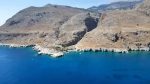 Vista panorámica de una bahía griega rodeada por colinas áridas bajo un sol intenso.