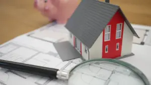 Maqueta de casa, lupa y hucha sobre planos de arquitecto para analizar hipotecas.