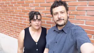 Un hombre y una mujer posan de pie frente a una pared de ladrillos rojos.